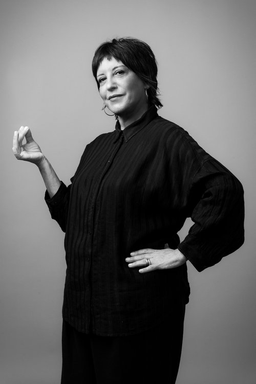Black and white portrait of Auriel Majumdar. She has short black hair with a short fringe. She is wearing a black shirt dress and is clicking her fingers whilst looking at the camera.