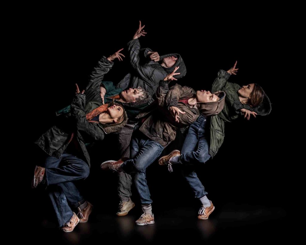 5 dancers wearing coats and jeans lean back in a arms crossed pose.