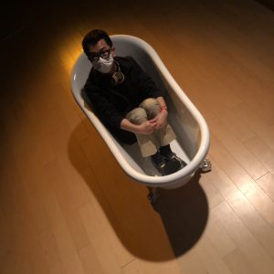 Abel photographed from above. He is sat fully clothed in a bathtub and looking up at the camera whilst wearing a surgical mask.
