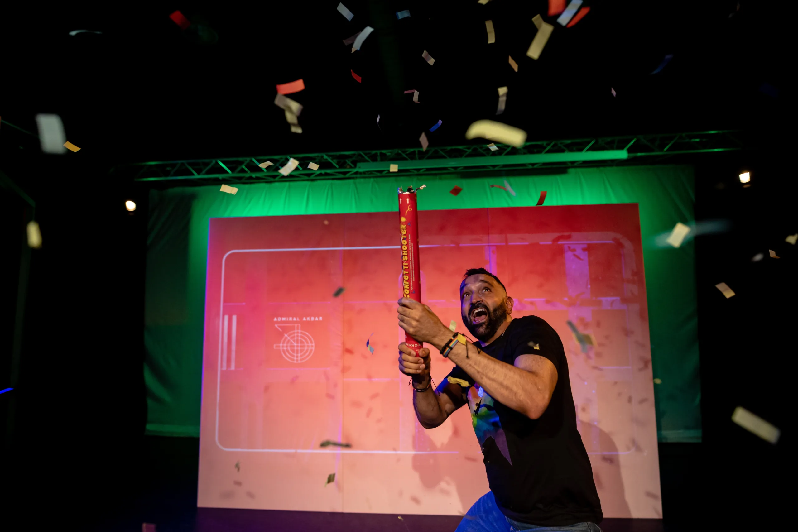 A brown man in a black t-shirt and with a neatly trimmed black beard lets off a confetti cannon on stage.
