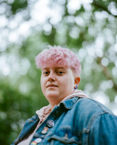 Daisy is stood in front of a tree which is out of focus and they have short pink hair and is wearing a blue denim jacket.