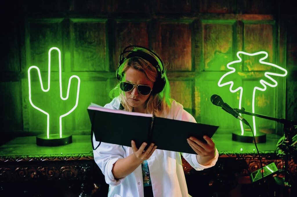 Fran Bundy looks in a book. She is wearing headphones and sunglasses. The room is dark and illuminated by green cactus and palm tree neon lights.