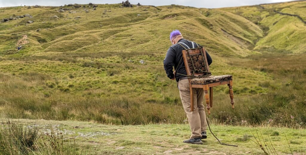 A person walking up the moors with a heavy chair strapped to his back