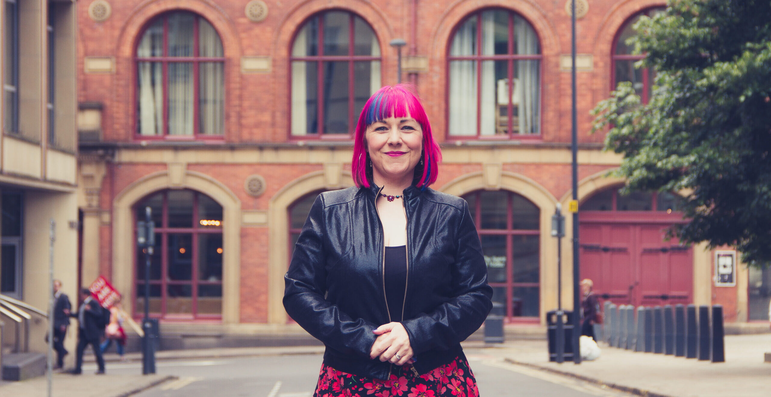 Peg Alexander stands on an urban street and has pink and purple hair and wears a leather-look jacket