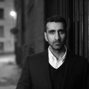 A black and white photo of Amit who is a south Asian male with short dark hair and is standing on a period street scene