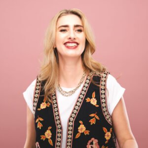 Jessica Knappett is a white woman with long blonde hair. She's stood in front of a pink background and smiles approachably to someone just off camera
