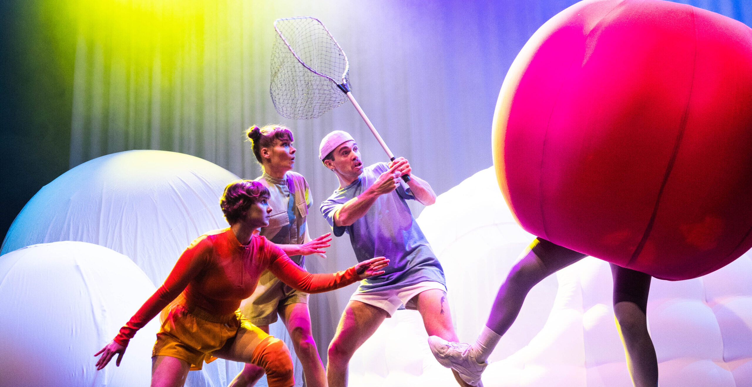 A cast of 3 people hold a net as if to try catch another cast member wearing an oversized red fruit costume
