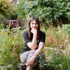 Julia is a white woman with long brown hair. She sits in a grassy woodland landscapa