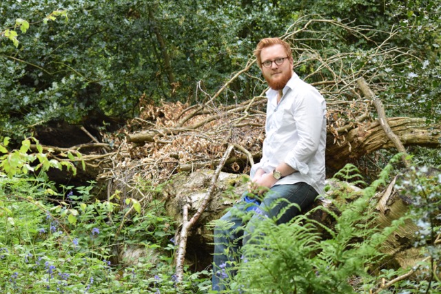 Photo of Peter Reed, a white man in his mid thirties. Short ginger hear and beard with glasses. He is sitting on a fallen tree amongst woodland.