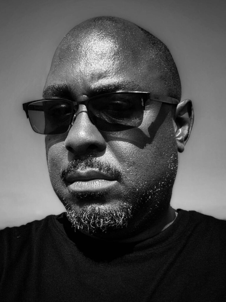 Headshot of Shaun Connell, a Black British man in sunglasses and a black t-shirt, taken on a Caribbean cruise with the sky as the backdrop.