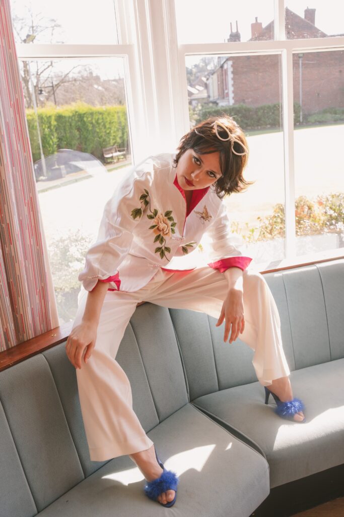 Hannah Hu sitting in a white suit by a window with sunglasses and fluffy heels.