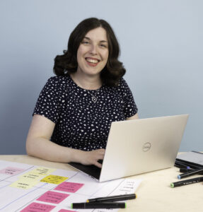 Photo of Sophie Greenwood, a white woman in her late-thirties. She has long brown curly hair and is wearing a blue spotty dress. The photo shows Sophie working at her laptop, surrounded by sticky notes and pens.