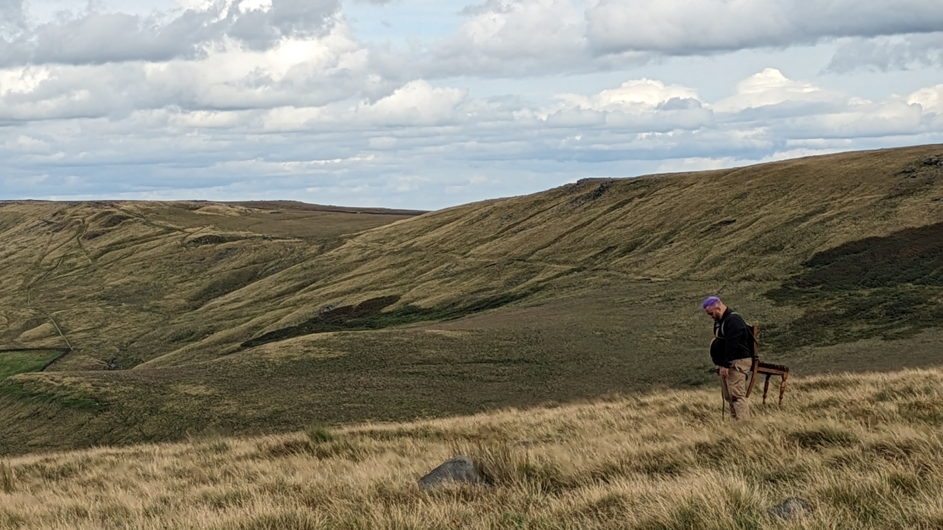 A still from the Bradford Producing Hub film still 'Held by the Wind' - an Empowered cohort commission. The scene shows the rolling hills surrounding Todmorden, with artist Chris Cambell visible with his head bowed, and an antique chair strapped to his back. 