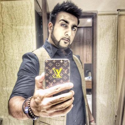 Davinder is a South Asian male. He his holding a phone with a Louis Vuitton case to take a selfie in the mirror. He has short dark brown hair and a short beard. He is wearing a dark blue shirt with a brown waistcoat.