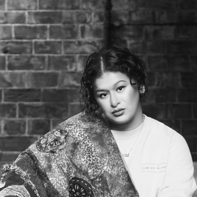 A black and white photograph of Aamta, she is looking to camera and is wearing a white sweater and has a appliqued cloth draped over her right shoulder. Aamta's hair is dark and curly and tied back with a centre parting with loose curls framing her face.